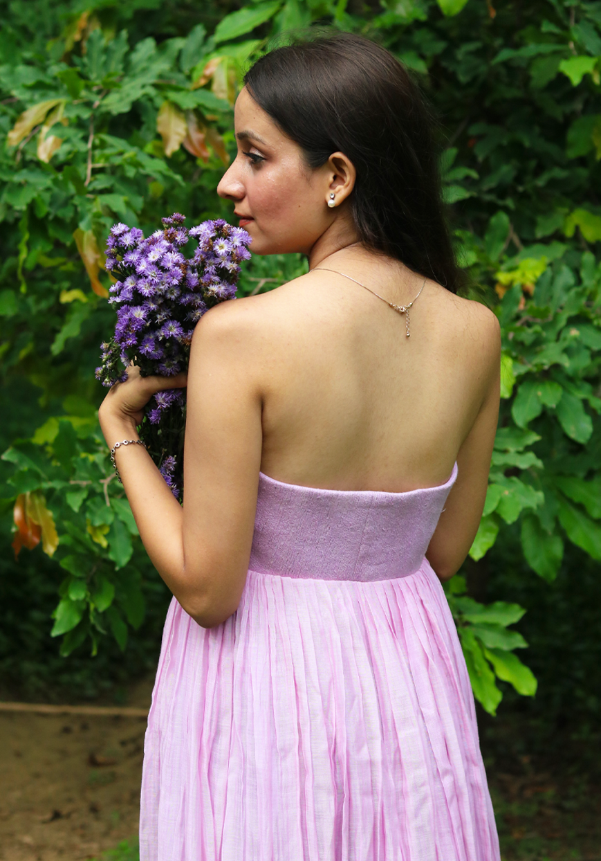 A woman wearing a strapless light pink pleated dress stands outdoors in front of green foliage, holding a bouquet of purple flowers.