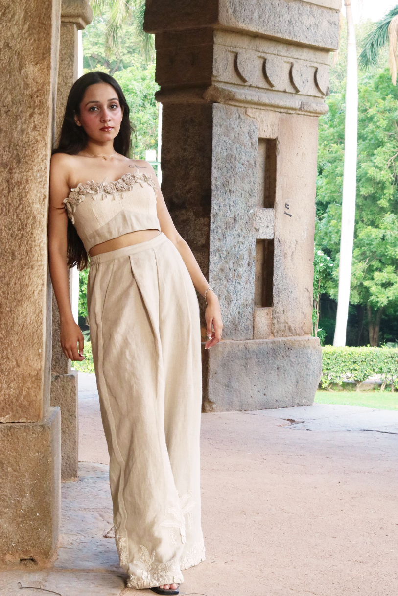 A woman wearing a beige strapless crop top with floral applique and matching high-waisted bottoms, standing in a stone corridor with greenery visible in the background.