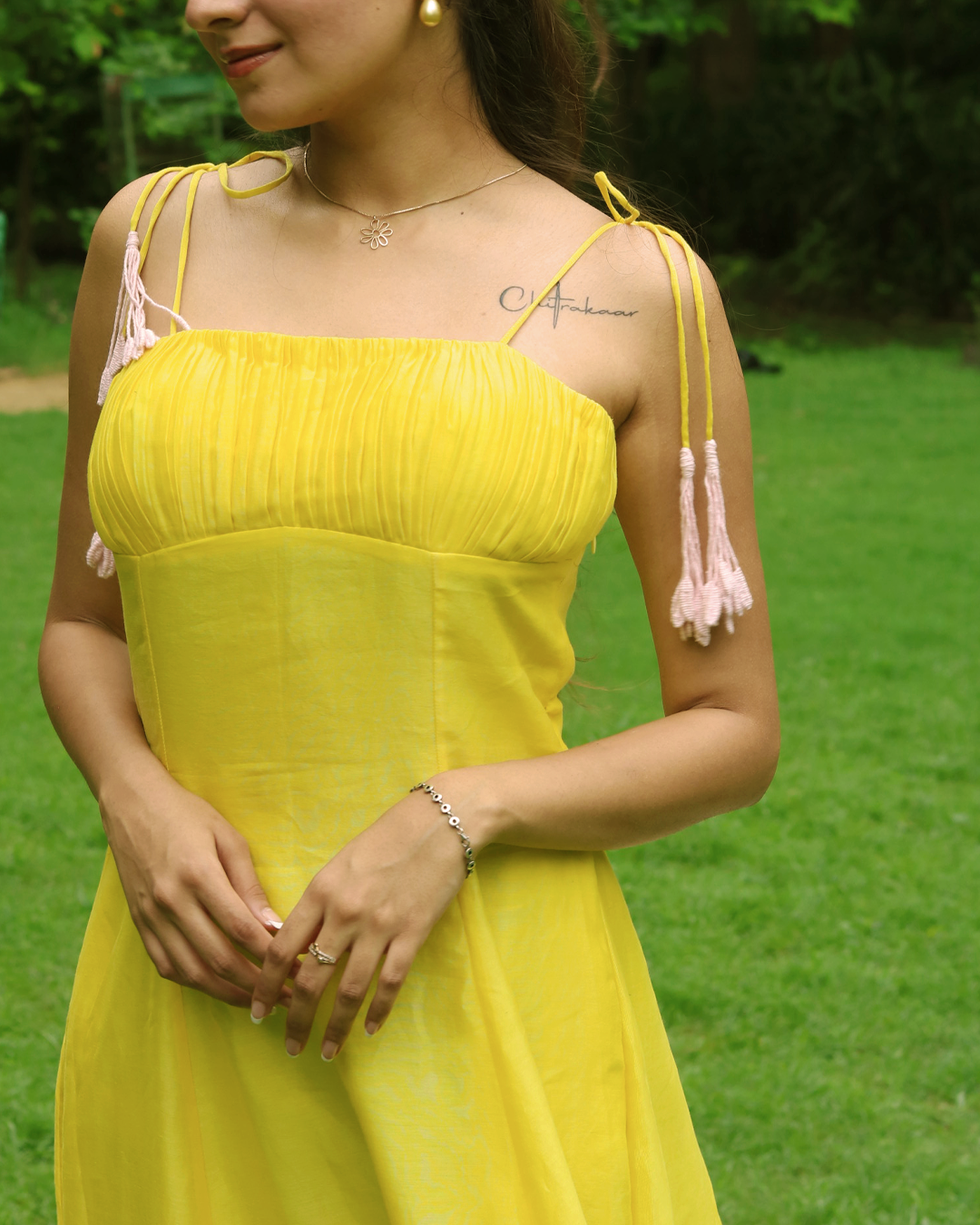 A woman wearing a bright yellow sleeveless dress with light pink tassel shoulder straps, standing outdoors on a green lawn.