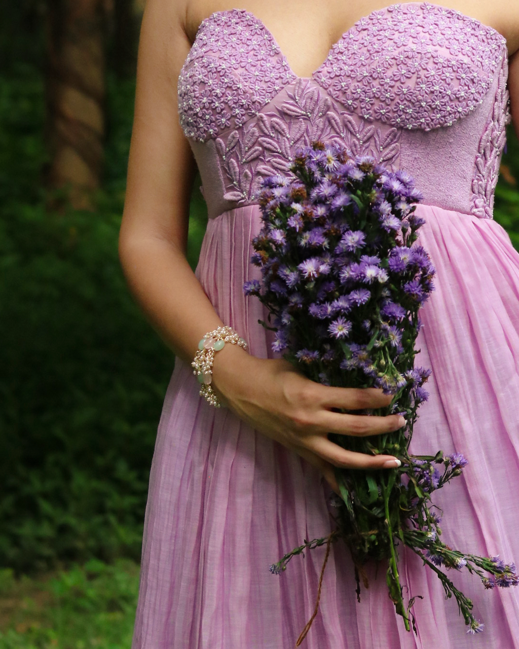 A woman outdoors wearing a lavender strapless dress with a textured lace bodice and a flowing pleated skirt, holding a bouquet of purple flowers.