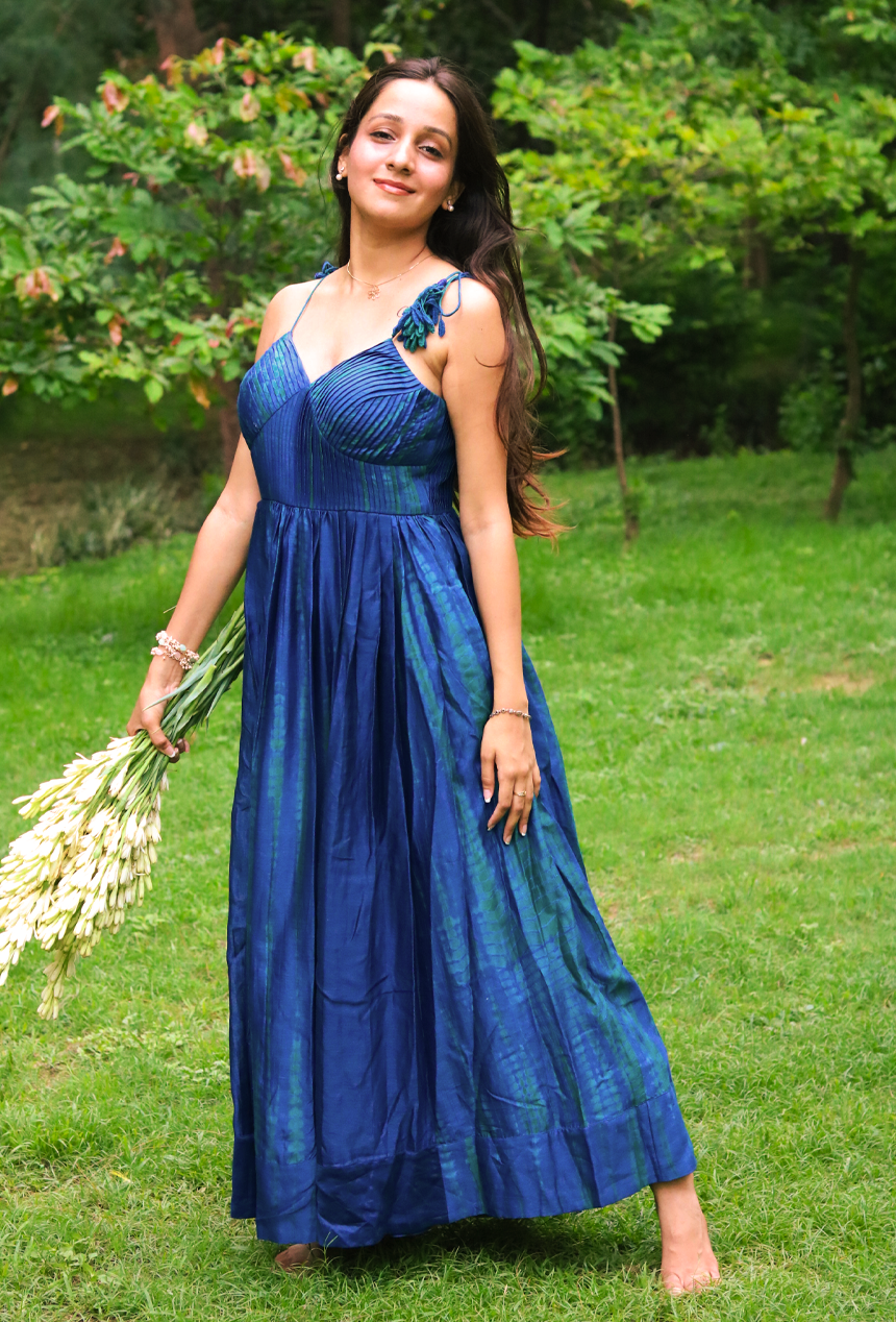A woman stands barefoot on a grassy lawn wearing a blue green tie dye sleeveless maxi dress. She is holding a bouquet of white flowers, with trees in the background.