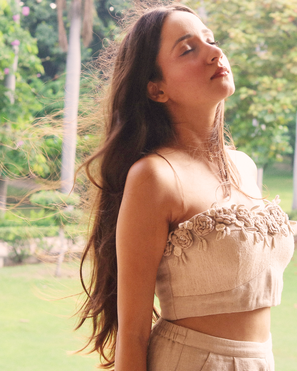 A woman outdoors in a park wearing a beige linen-style two-piece set with a strapless cropped top featuring 3D floral embroidery and matching high-waisted pants.