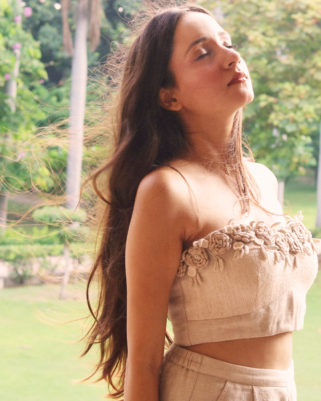 A woman outdoors in a park wearing a beige linen-style two-piece set with a strapless cropped top featuring 3D floral embroidery and matching high-waisted pants.