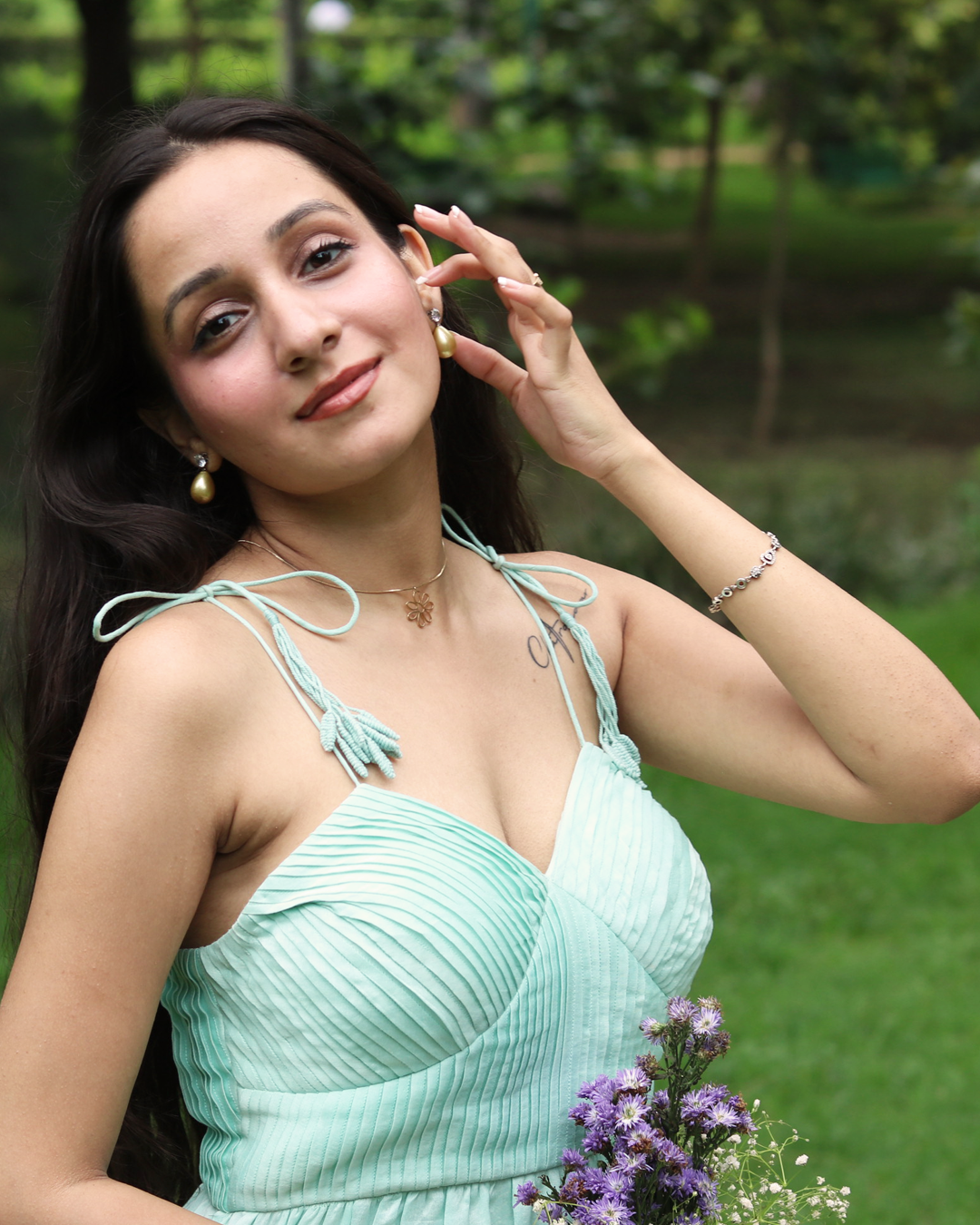 A woman wearing a sea blue sleeveless maxi dress with embroidered palm tree motifs on the skirt, photographed outdoors in a park setting.