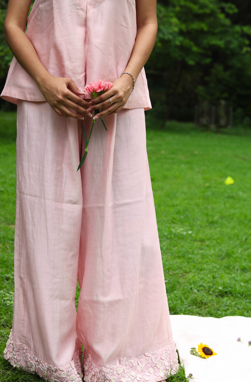 A person outdoors wearing a light pink two-piece outfit with a sleeveless top and wide-leg pants featuring lace trim at the hems, standing on a grassy lawn and holding a pink flower.