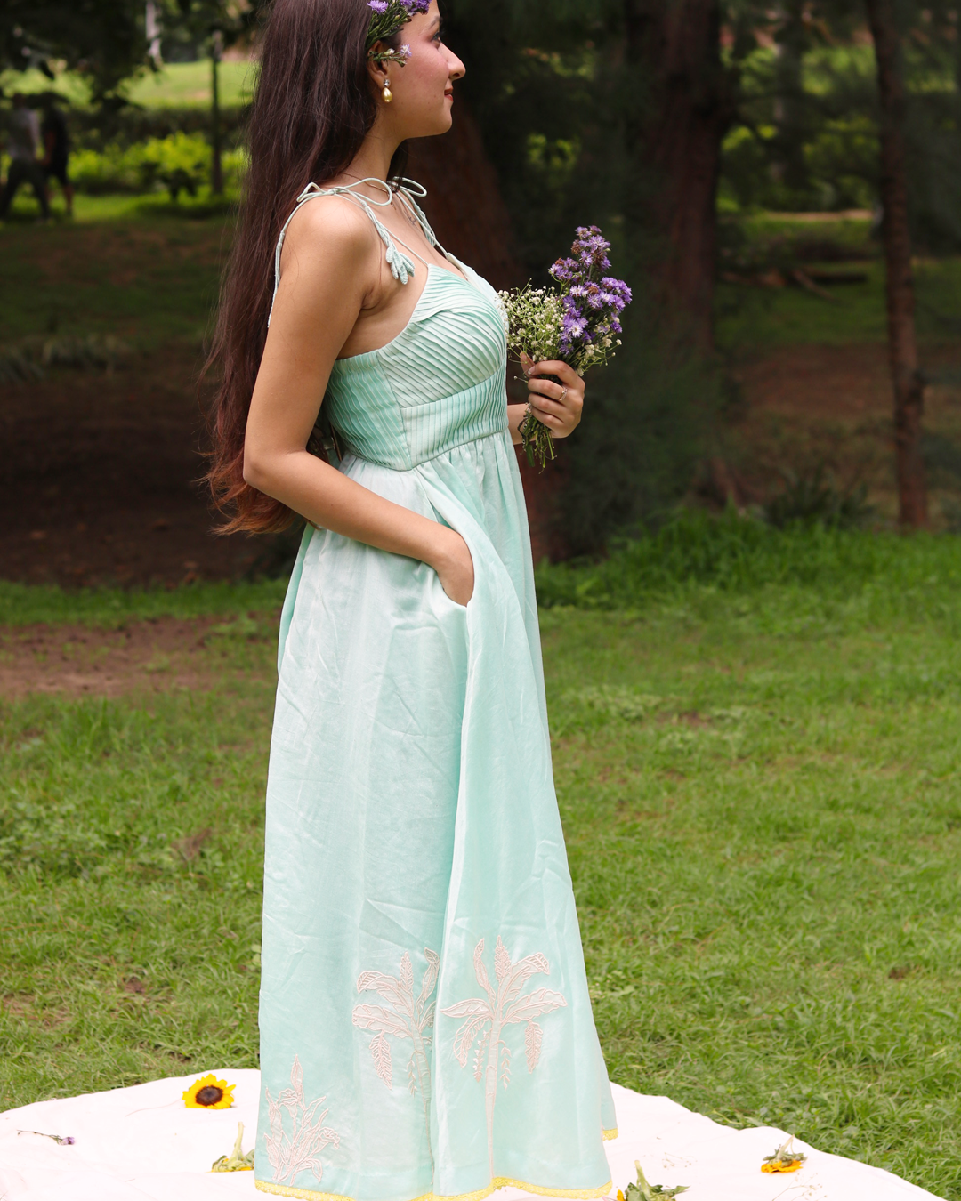 A woman wearing a sea blue sleeveless maxi dress with embroidered palm tree motifs on the skirt, photographed outdoors in a park setting.