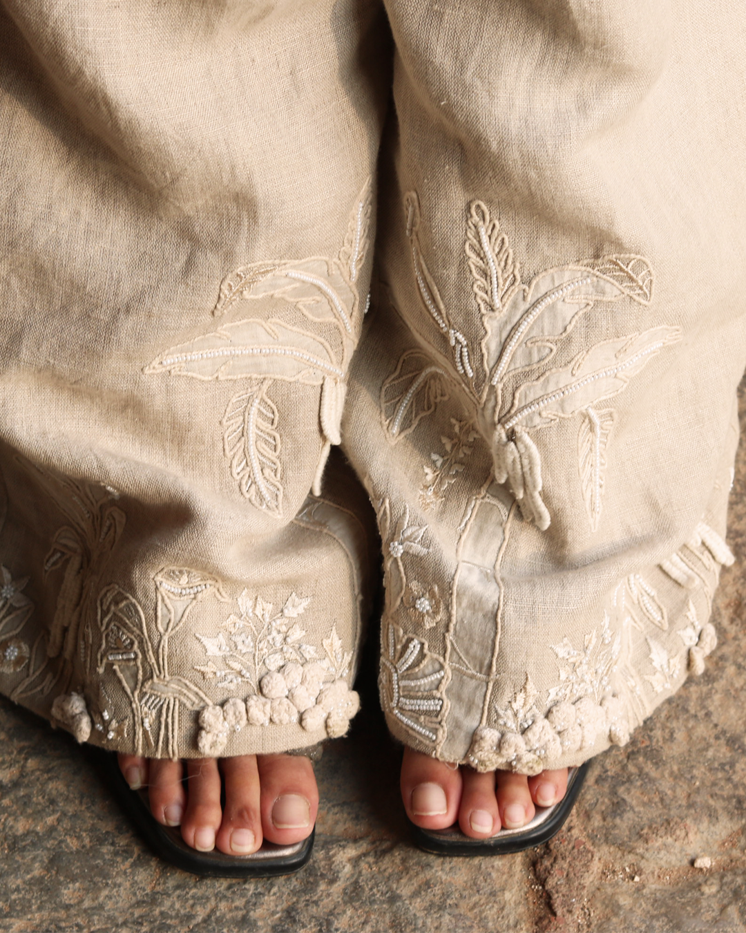 Close-up of a person wearing beige linen pants with intricate white floral embroidery and beadwork along the wide hems, paired with black slide sandals on a stone surface.
