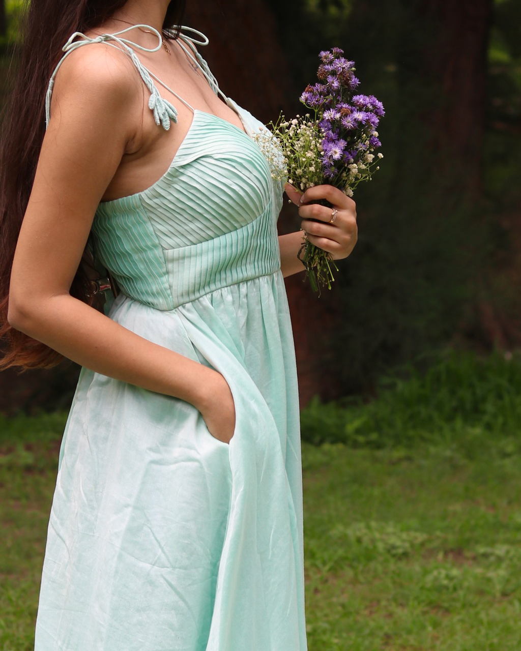 A woman wearing a sea blue sleeveless maxi dress with embroidered palm tree motifs on the skirt, photographed outdoors in a park setting.