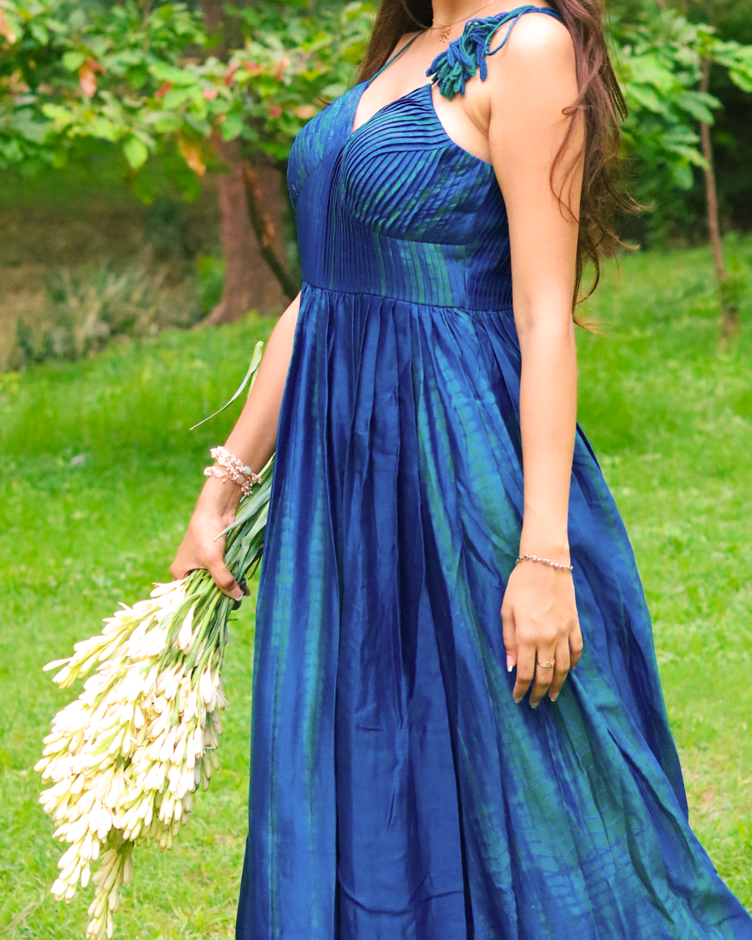 A woman stands barefoot on a grassy lawn wearing a blue green tie dye sleeveless maxi dress. She is holding a bouquet of white flowers, with trees in the background.