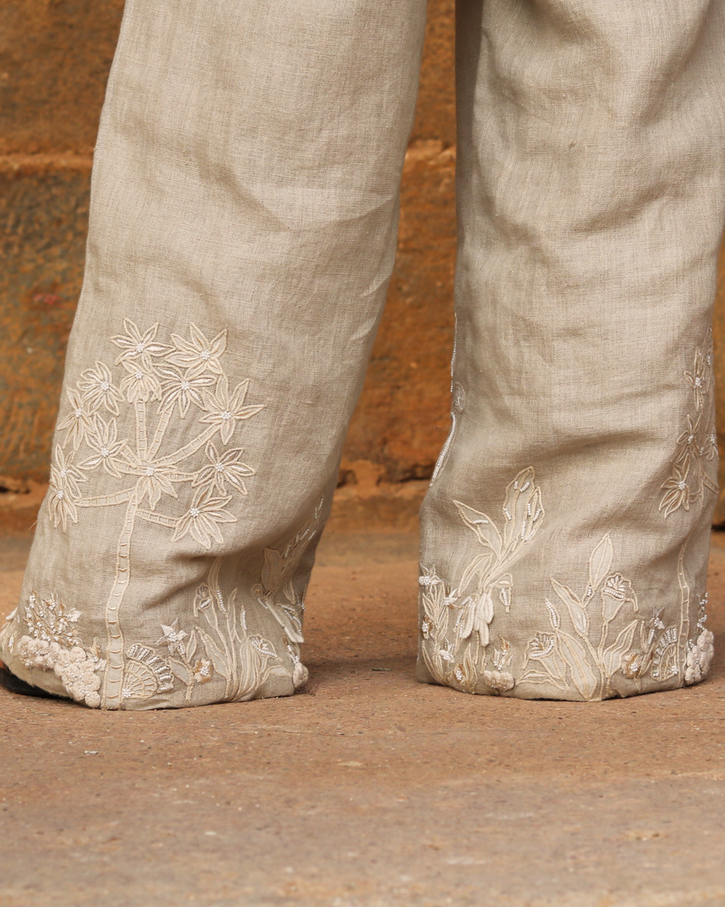 Close-up of a person's legs wearing wide-leg linen pants in a light beige color, featuring detailed floral embroidery near the hems, standing on a rustic stone surface.