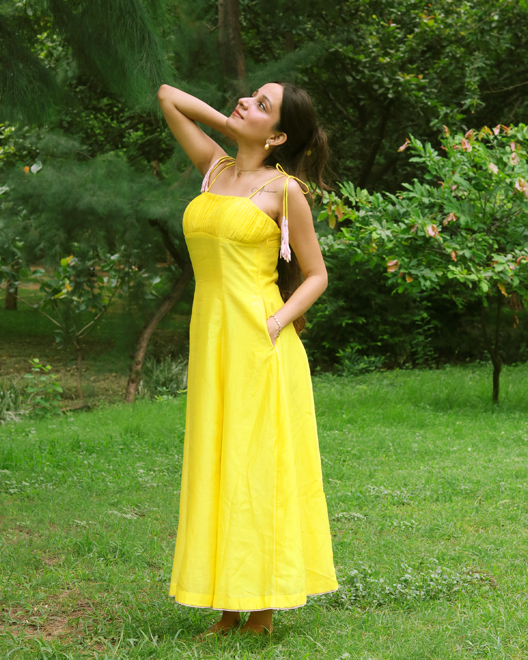 A woman wearing a bright yellow strapless maxi dress with white tassel details on the straps, standing in a grassy park with trees in the background.