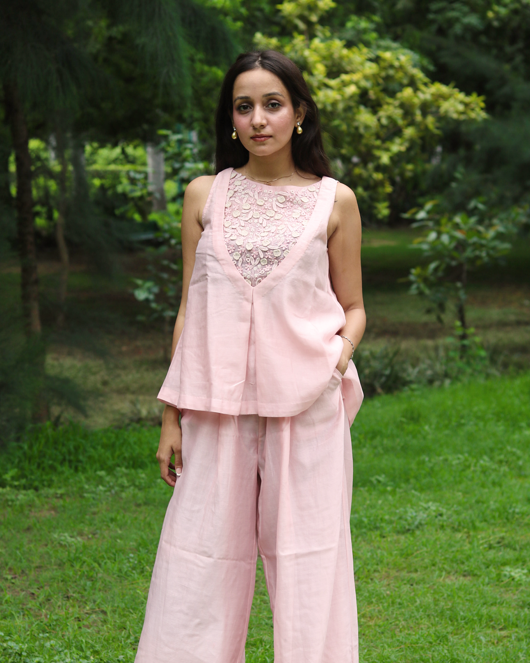 A woman stands outdoors on a grassy lawn wearing a light pink sleeveless linen top with a floral lace V-neckline and matching wide-leg trousers.