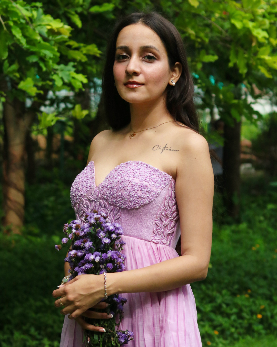A woman wearing a strapless lavender dress stands outdoors in a garden, holding a bouquet of purple flowers. The dress has a textured, lace-like sweetheart bodice and a pleated skirt.
