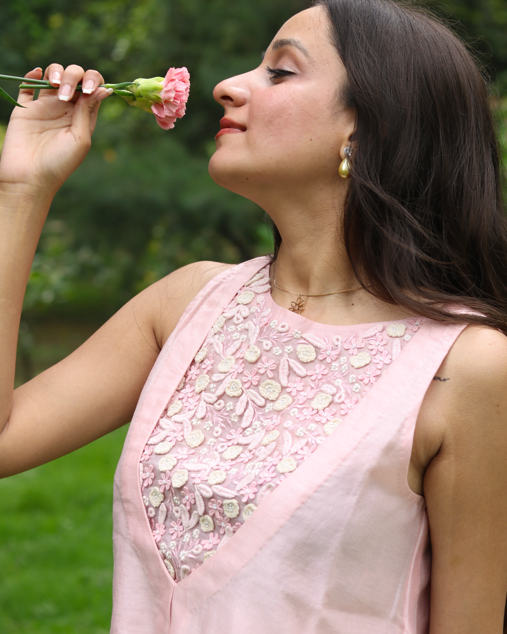 A woman in a sleeveless light pink dress with a heavily embroidered floral neckline, holding a pink carnation flower near her face against a blurred green outdoor background.