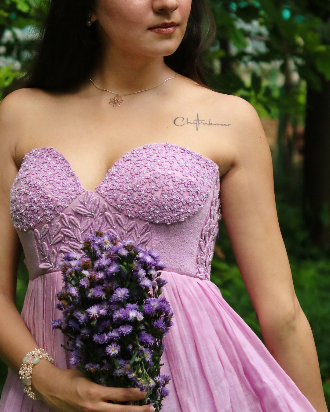 A woman outdoors wearing a strapless lavender dress with a textured floral bodice and a flowing tulle skirt, holding a bouquet of purple flowers. A small script tattoo is visible on her upper chest.
