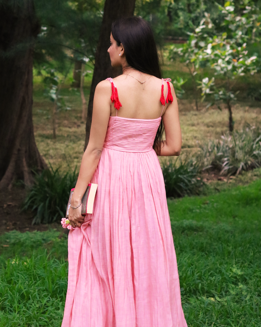 A woman wearing a pink pleated maxi dress with red tassel shoulder straps, photographed from behind in an outdoor park setting.