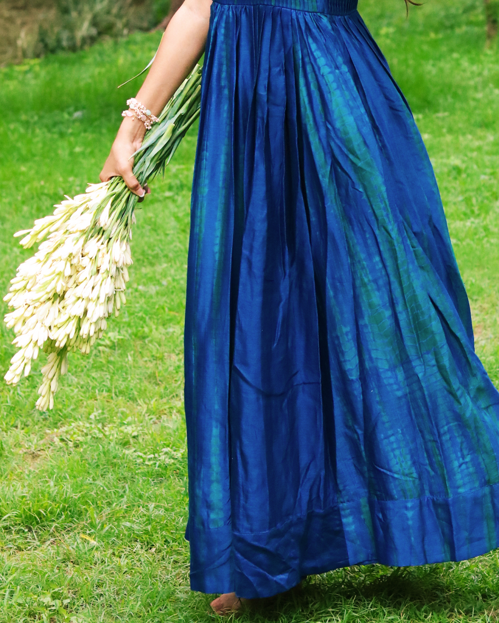 A woman stands barefoot on a grassy lawn wearing a blue green tie dye sleeveless maxi dress. She is holding a bouquet of white flowers, with trees in the background.