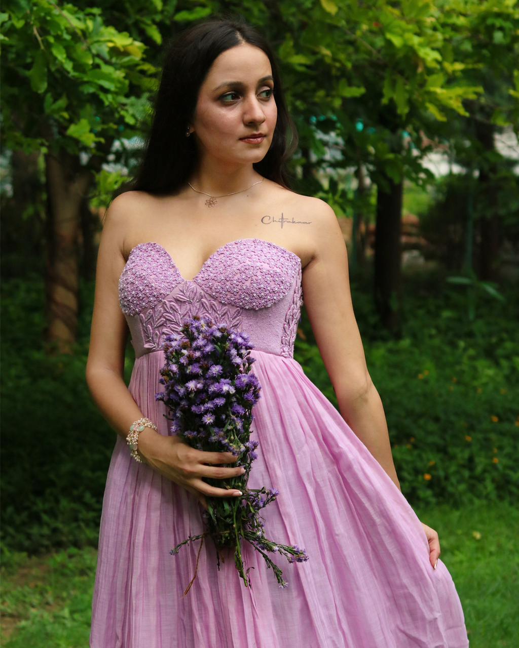 A woman wearing a strapless lavender-pink gown with a textured lace bodice and a flowing tulle skirt, holding a bouquet of purple flowers in a garden setting.