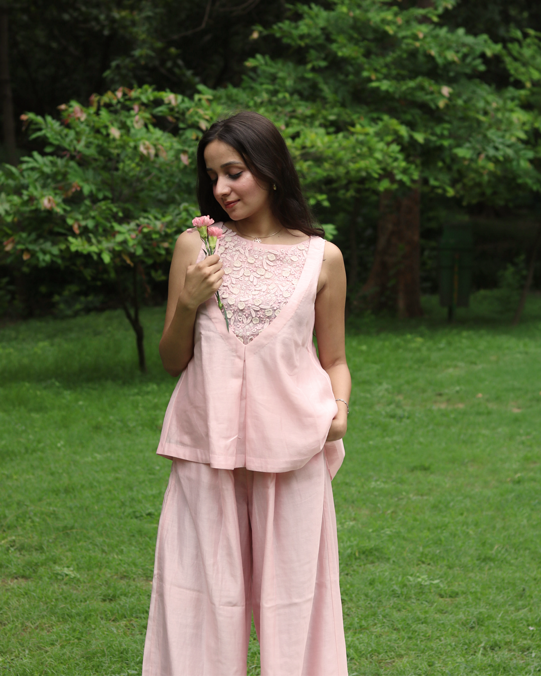 A woman stands outdoors on a grassy lawn wearing a light pink sleeveless linen top with a floral lace V-neckline and matching wide-leg trousers.