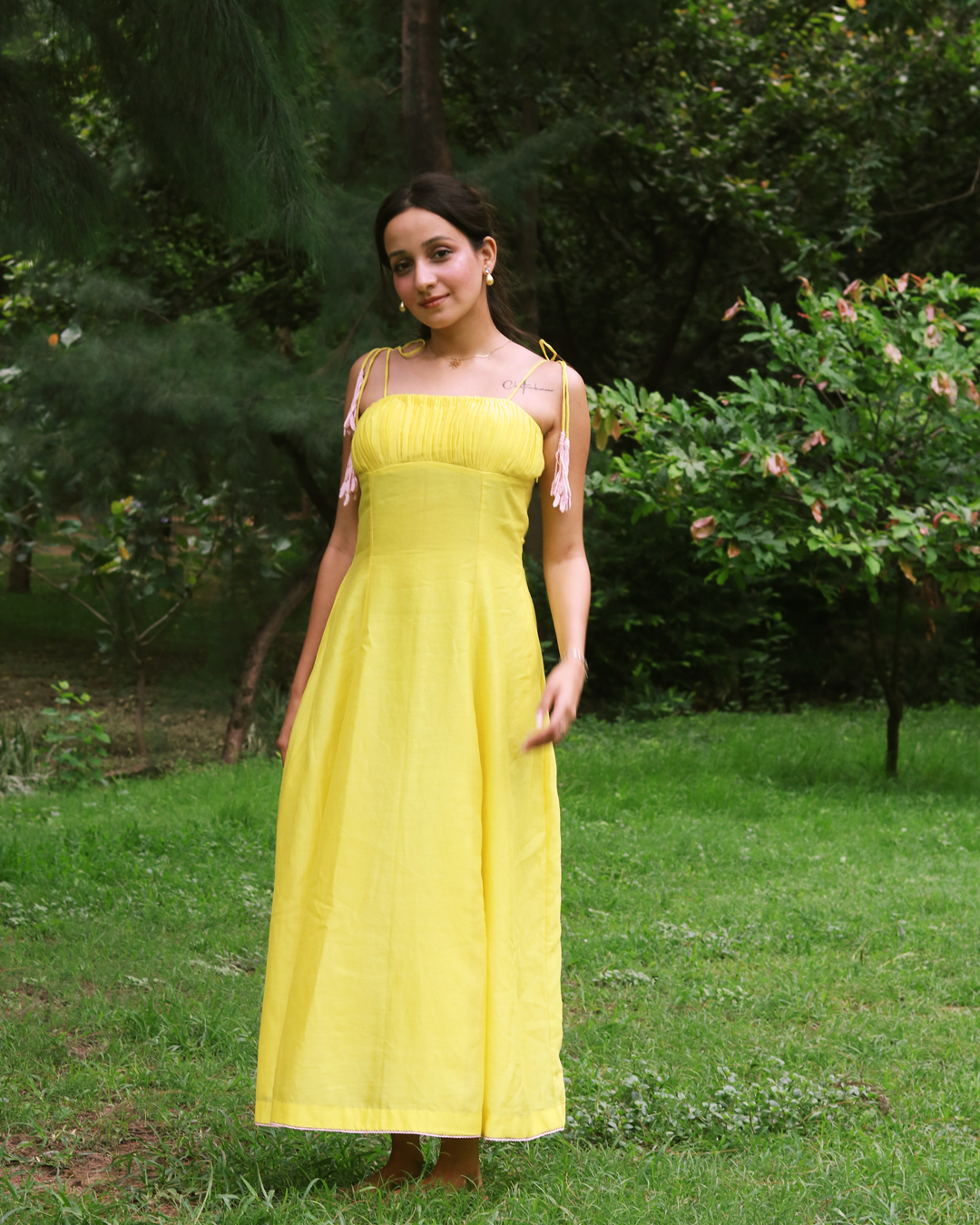A woman wearing a bright yellow sleeveless maxi dress with pink ribbon tie details on the straps, standing outdoors on a grassy lawn with trees in the background.