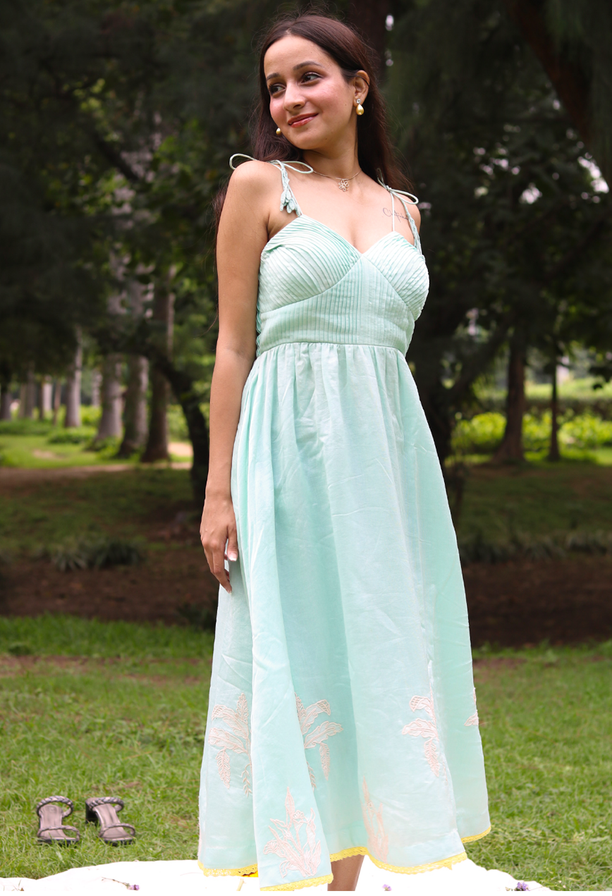A woman wearing a sea blue sleeveless maxi dress with embroidered palm tree motifs on the skirt, photographed outdoors in a park setting.