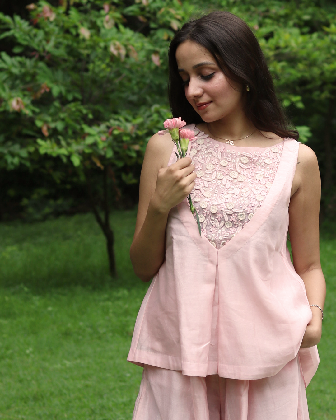 A woman stands outdoors on a grassy lawn wearing a light pink sleeveless linen top with a floral lace V-neckline and matching wide-leg trousers.