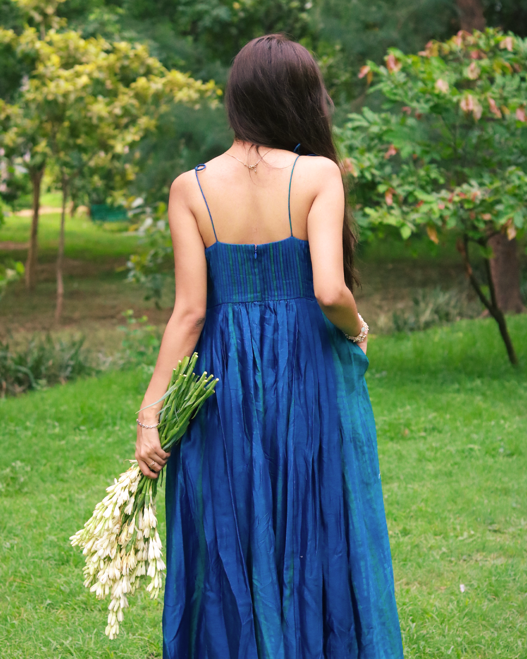A woman stands barefoot on a grassy lawn wearing a blue green tie dye sleeveless maxi dress. She is holding a bouquet of white flowers, with trees in the background.