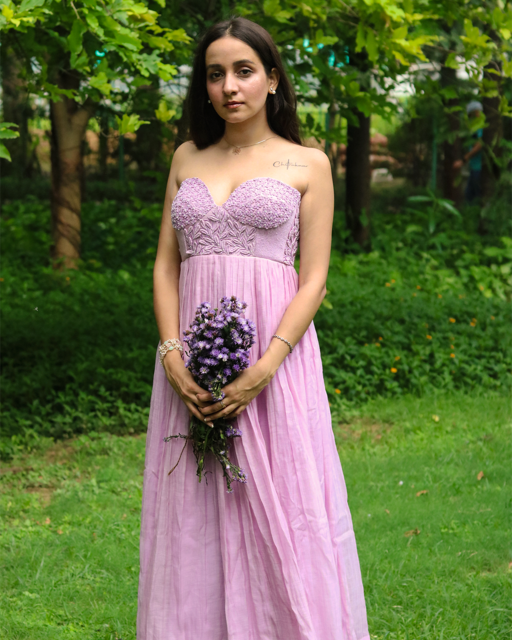 A woman stands outdoors in a lush green garden wearing a lavender strapless gown with a lace-embroidered bodice and a flowing tulle skirt, holding a small bouquet of purple flowers.