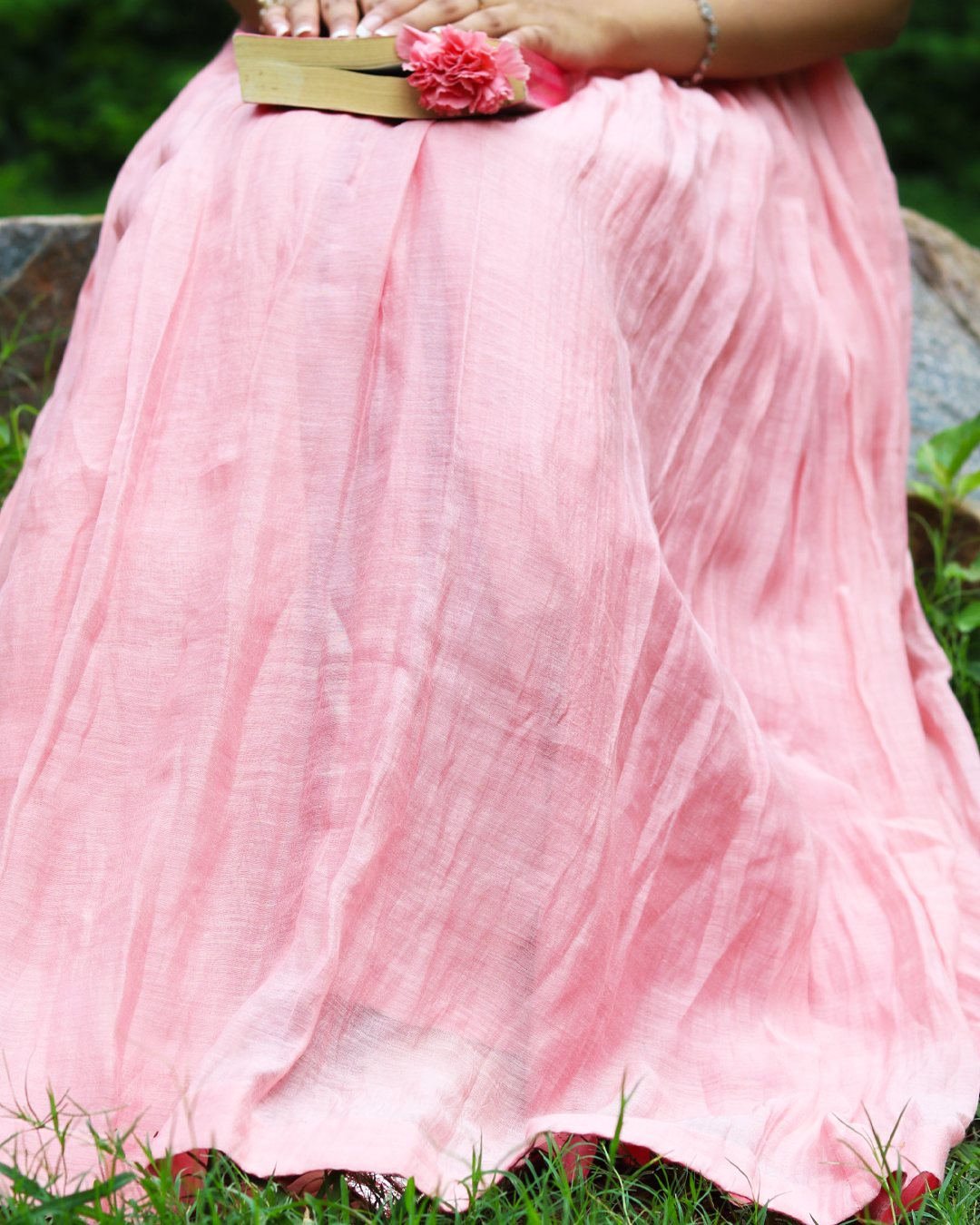 A close-up of a person wearing a flowing light pink skirt outdoors, seated on grass with a book and pink flower held near the waist.