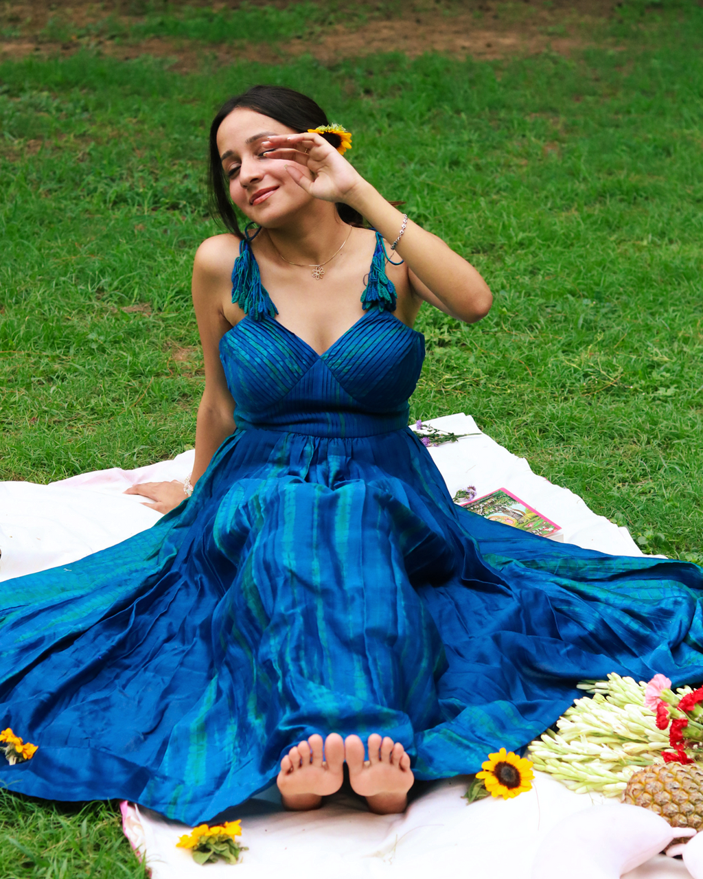 A woman sitting on a white blanket on a grassy lawn, wearing a flowing royal blue maxi dress with tie-shoulder straps and bare feet visible. Sunflowers and a basket of flowers are on the blanket beside her.