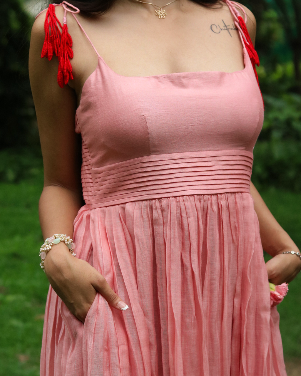 A woman wearing a dusty pink linen dress with red tassel shoulder ties, standing outdoors in a grassy garden setting.