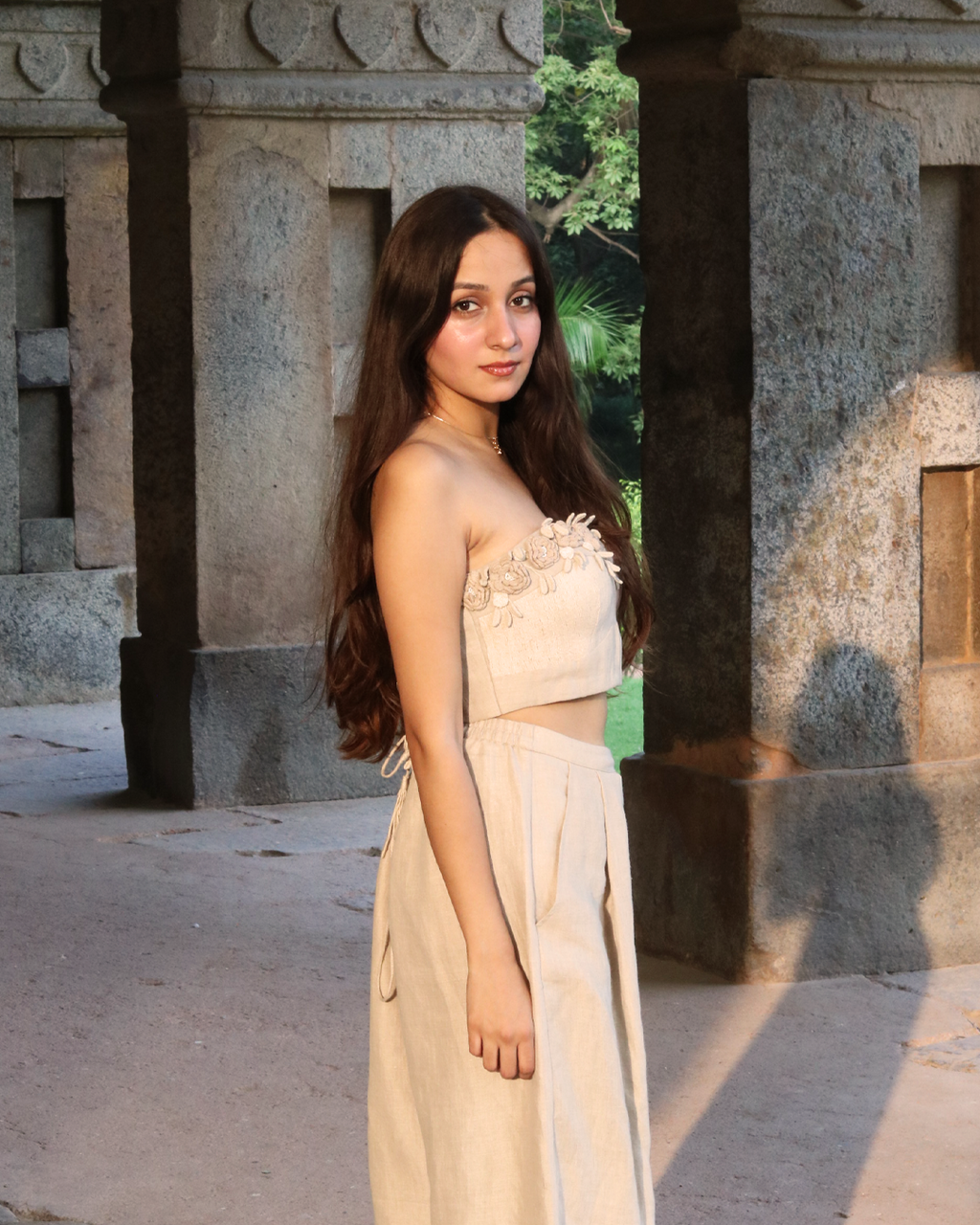 A woman wearing a beige strapless crop top with floral applique and matching high-waisted bottoms, standing in a stone corridor with greenery visible in the background.