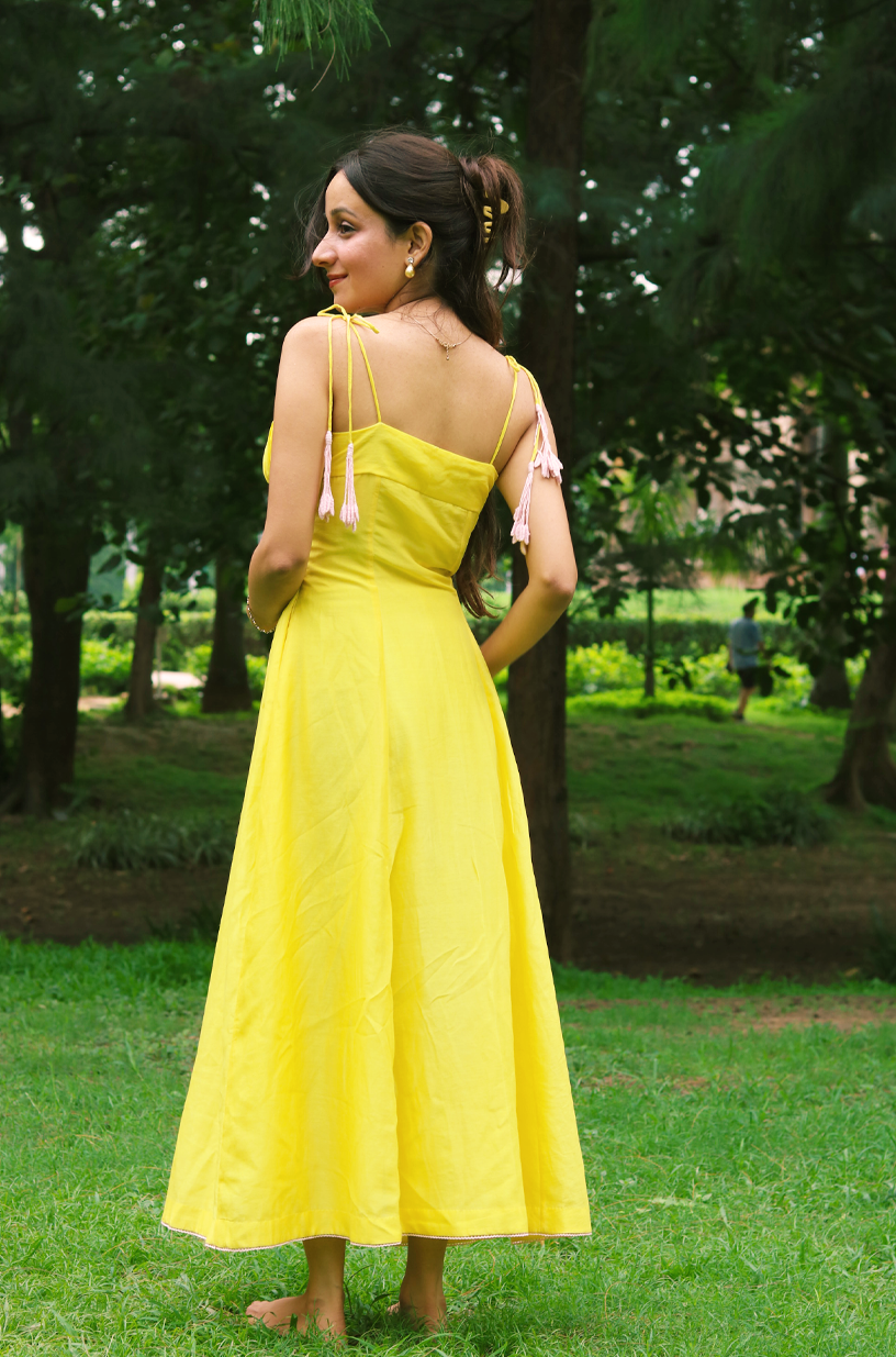 A woman stands barefoot in a grassy park wearing a bright yellow sleeveless maxi dress with thin shoulder ties and a fitted silhouette.