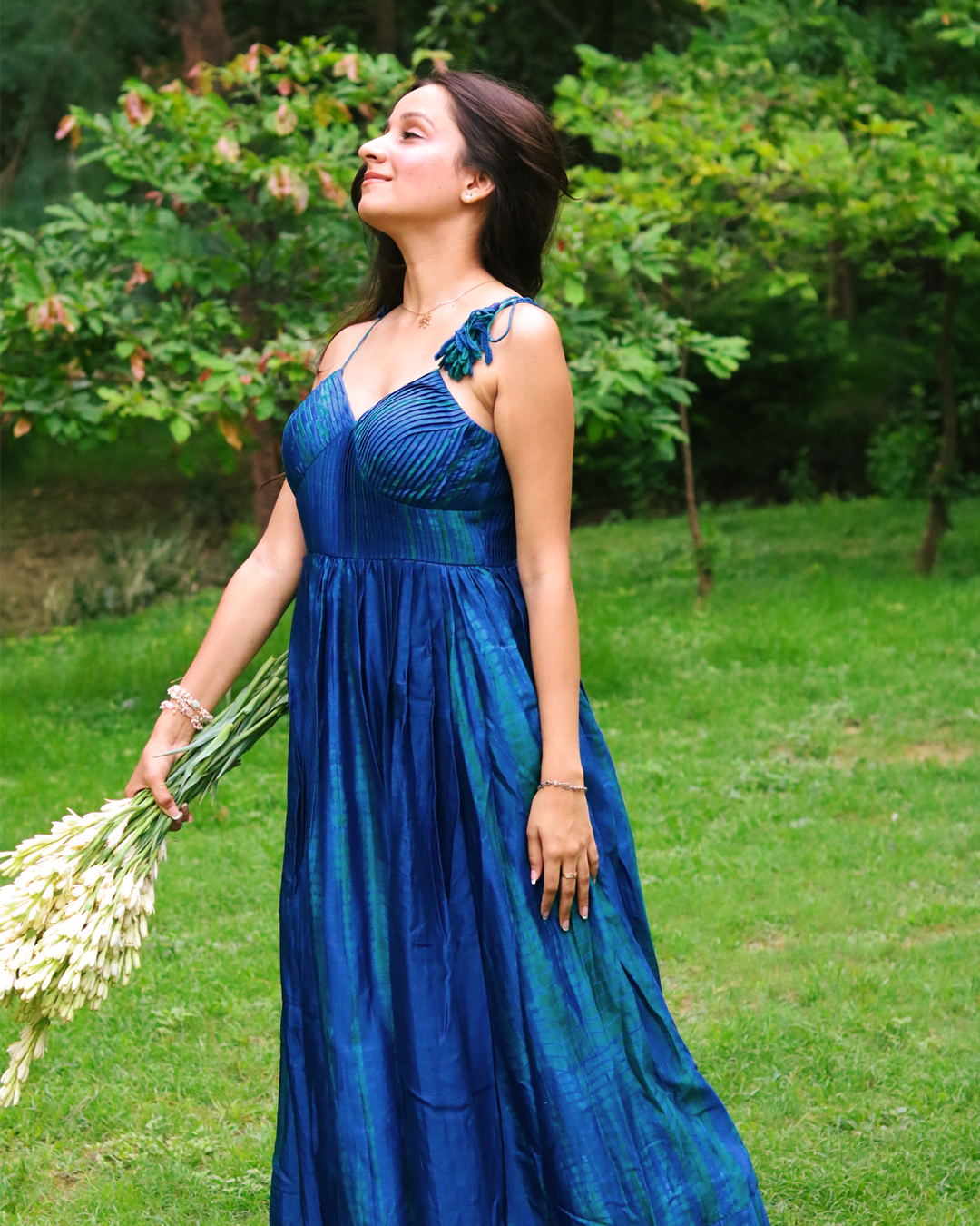 A woman stands barefoot on a grassy lawn wearing a blue green tie dye sleeveless maxi dress. She is holding a bouquet of white flowers, with trees in the background.