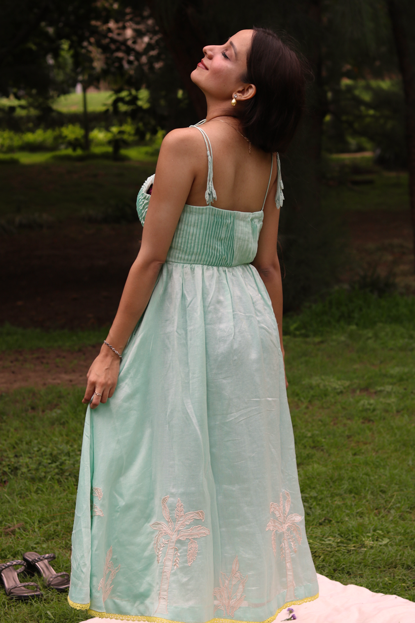 A woman wearing a sea blue sleeveless maxi dress with embroidered palm tree motifs on the skirt, photographed outdoors in a park setting.