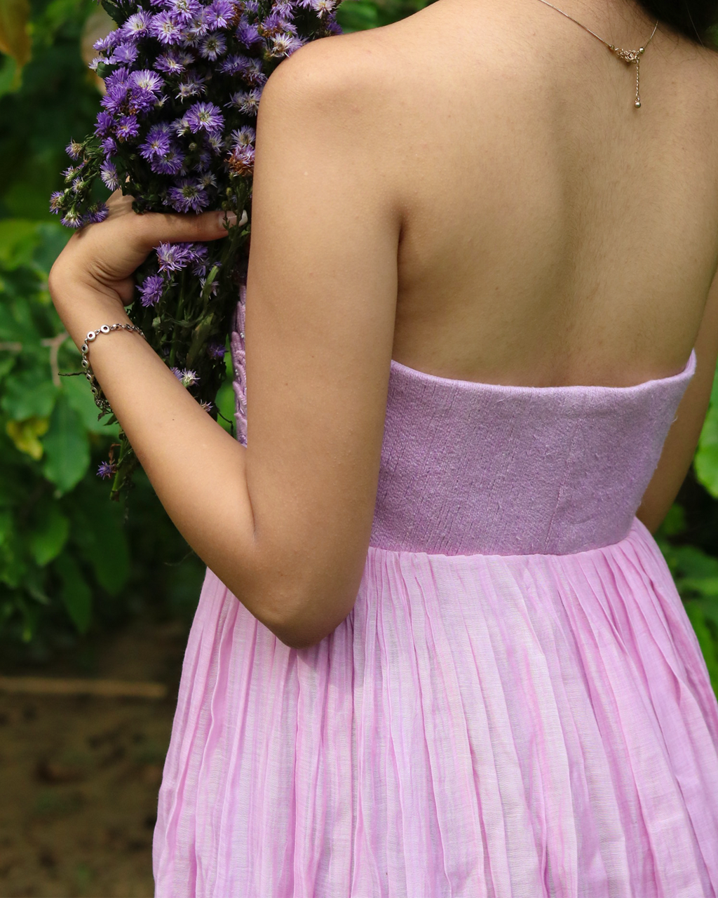 A woman shown from behind wearing a strapless light purple dress with a pleated skirt, holding a bouquet of purple flowers against a green garden background.