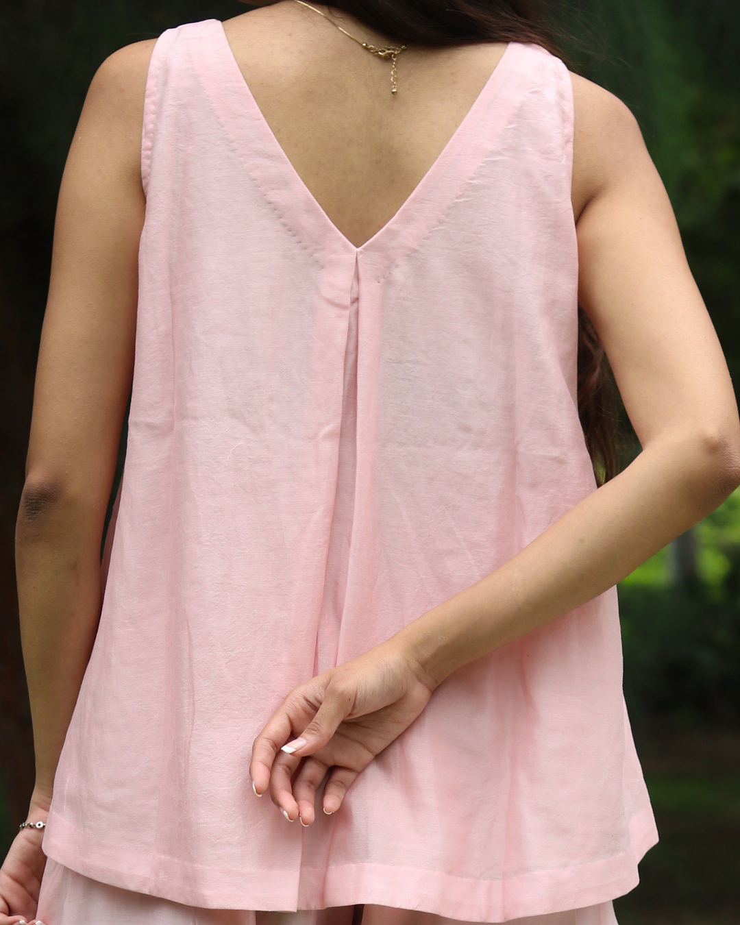 A woman wearing a light pink sleeveless linen top with a V-neckline, shown from the back in an outdoor setting with blurred green foliage in the background.
