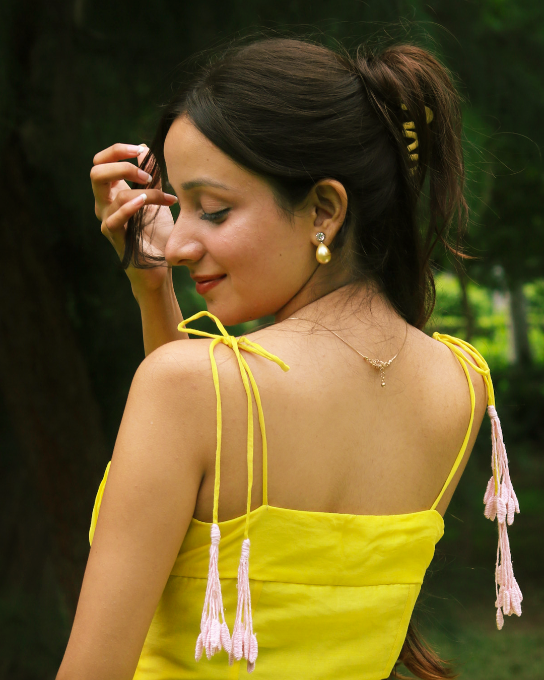 A woman wearing a bright yellow spaghetti-strap top with pink tassels hanging from the straps, photographed outdoors against a blurred green forest background.