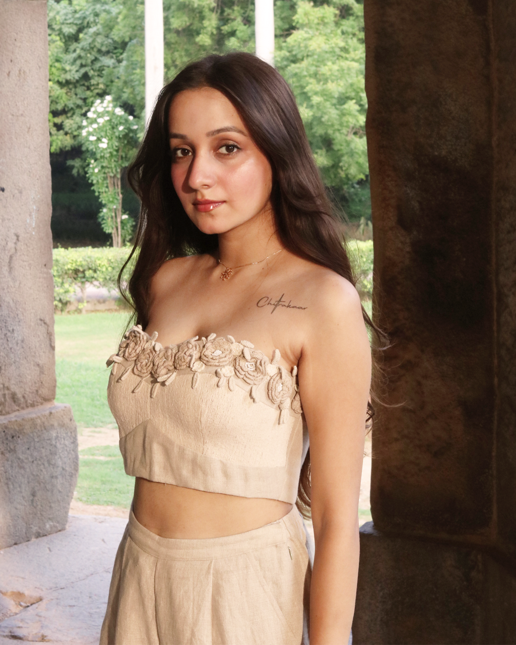 A woman wearing a beige strapless crop top with floral applique and matching high-waisted bottoms, standing in a stone corridor with greenery visible in the background.
