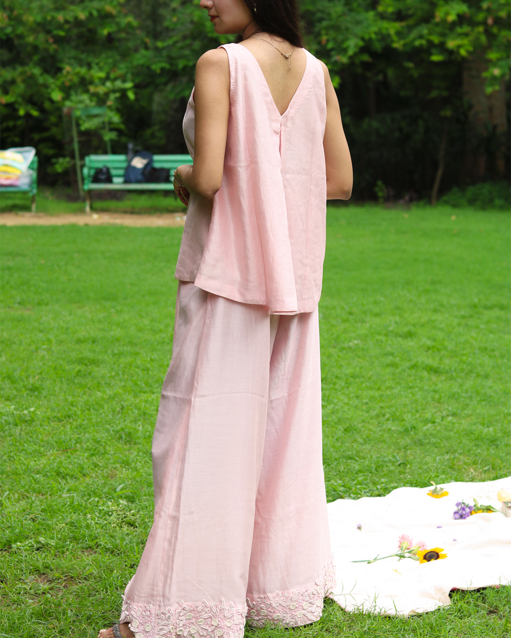 A woman stands outdoors on a grassy lawn wearing a light pink sleeveless linen top with a floral lace V-neckline and matching wide-leg trousers.