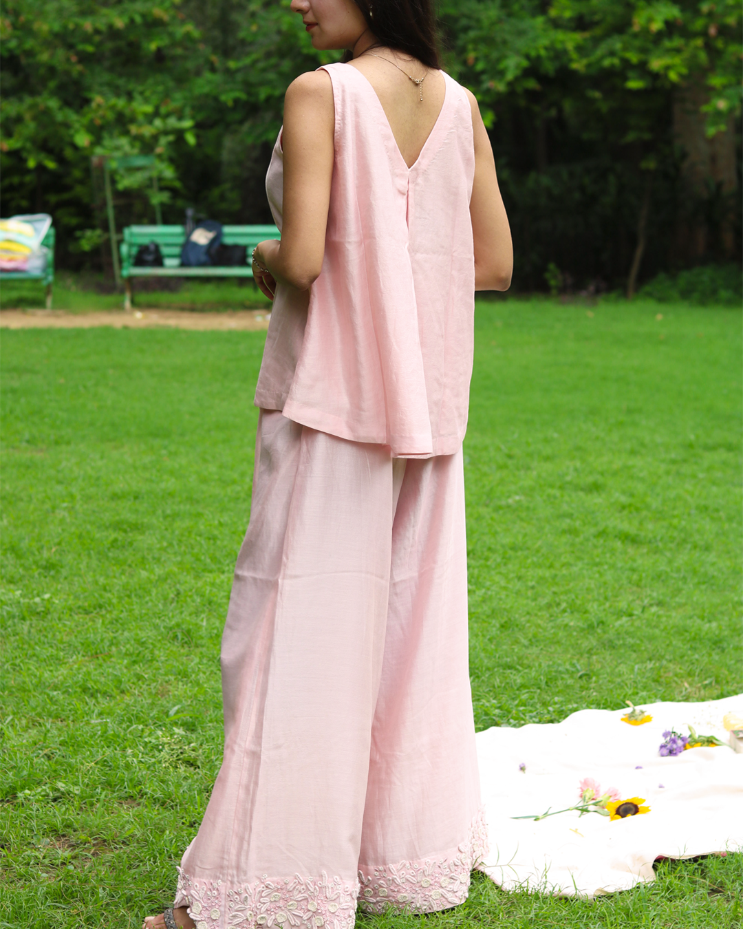A woman stands outdoors on a grassy lawn wearing a light pink sleeveless linen top with a floral lace V-neckline and matching wide-leg trousers.