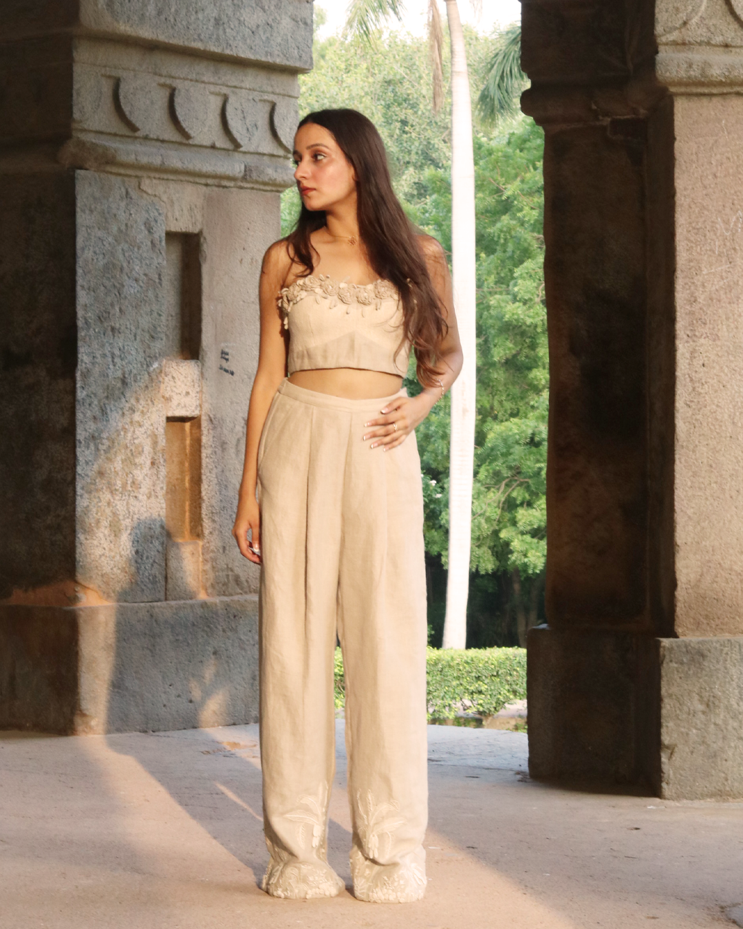 A woman wearing a beige strapless crop top with floral applique and matching high-waisted bottoms, standing in a stone corridor with greenery visible in the background.