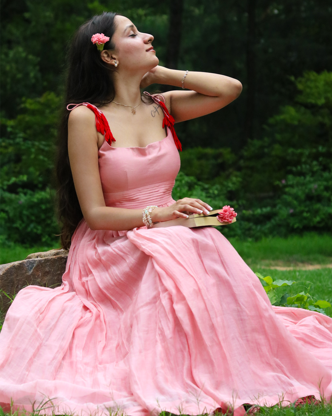 A woman in a flowing pink gown with red shoulder ties sits outdoors on a stone in a wooded setting, holding a small pink flower.