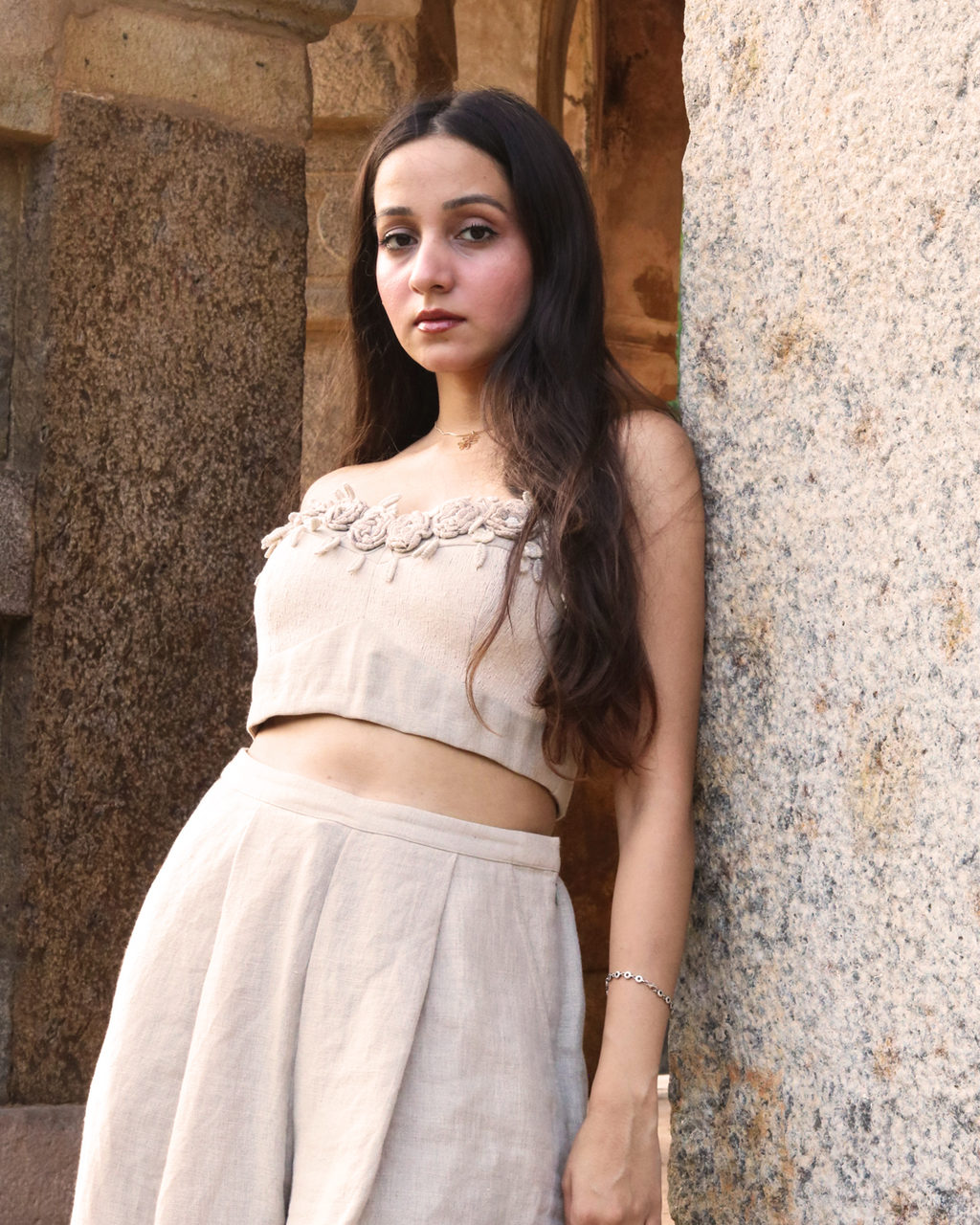 A woman wearing a beige strapless crop top with floral applique and matching high-waisted bottoms, standing in a stone corridor with greenery visible in the background.