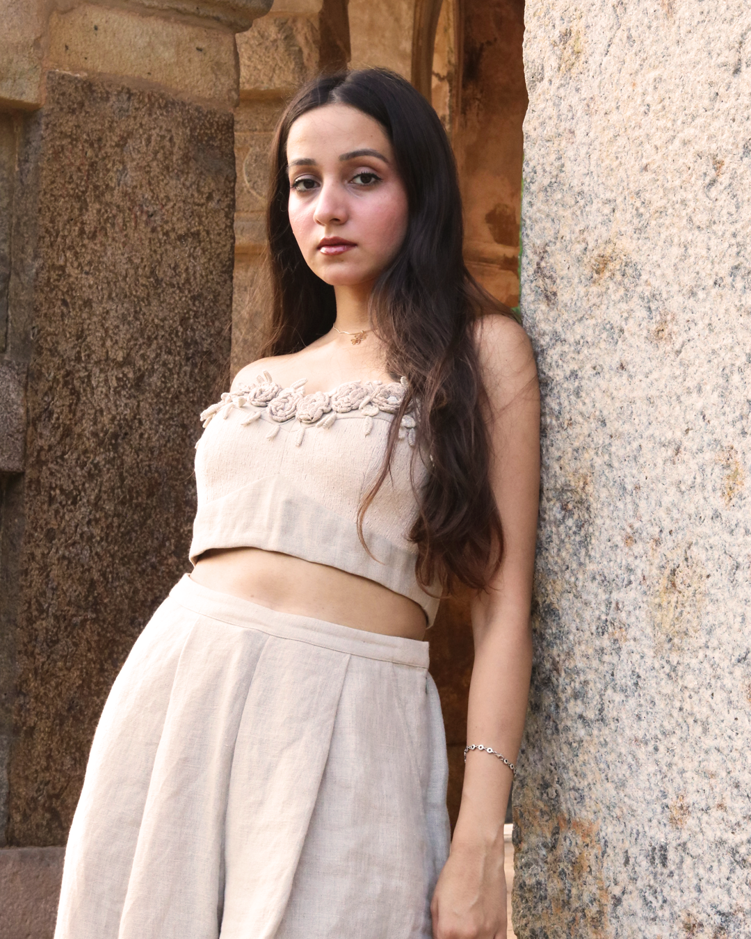 A woman wearing a beige strapless crop top with floral applique and matching high-waisted bottoms, standing in a stone corridor with greenery visible in the background.