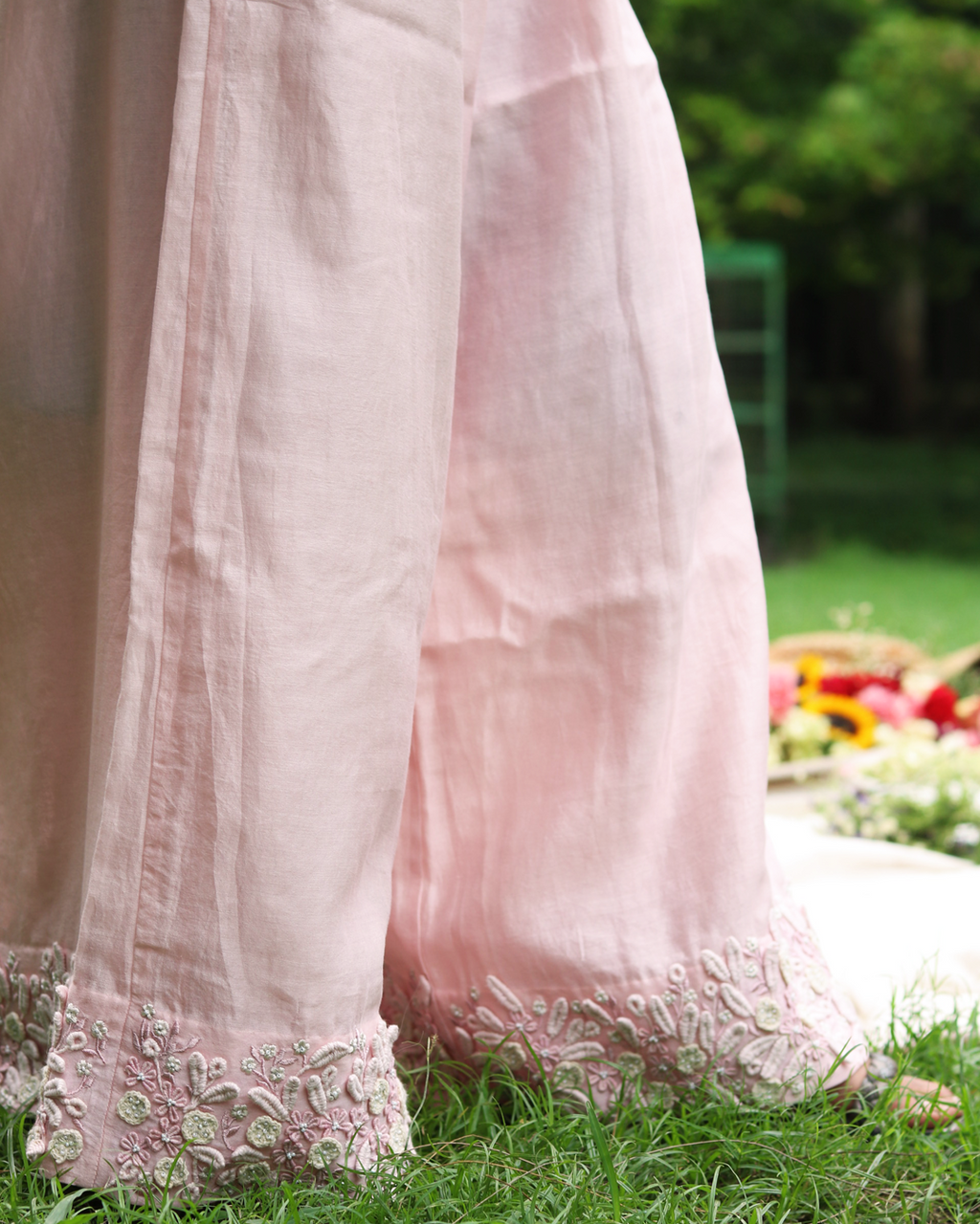 A close-up of a light pink traditional outfit with wide legs, featuring detailed floral embroidery along the hem, worn outdoors on a grassy lawn.