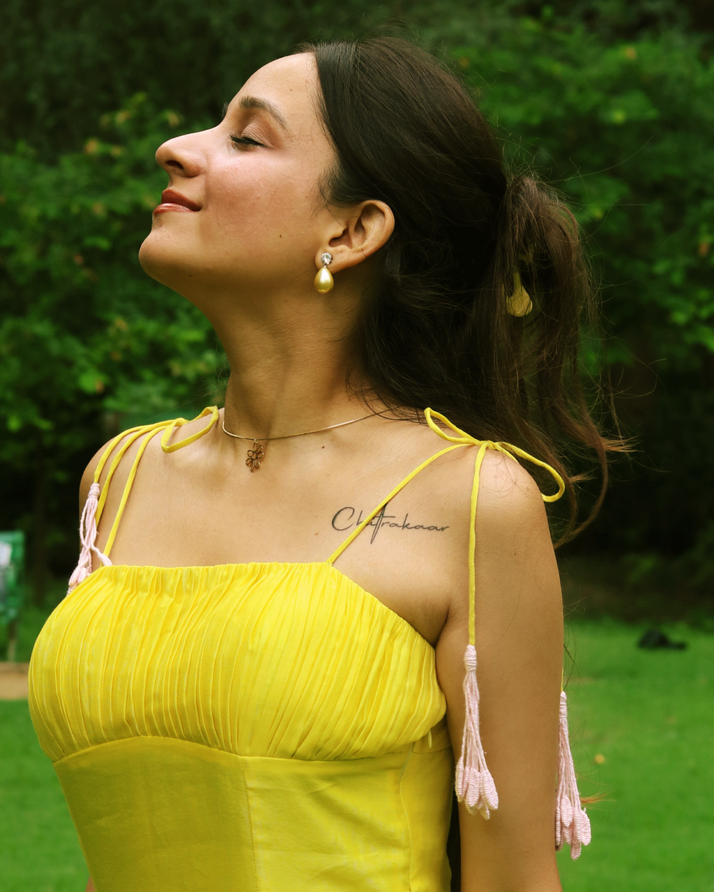 A woman wearing a bright yellow strapless dress with pink tassel shoulder ties, photographed outdoors against a green garden background.