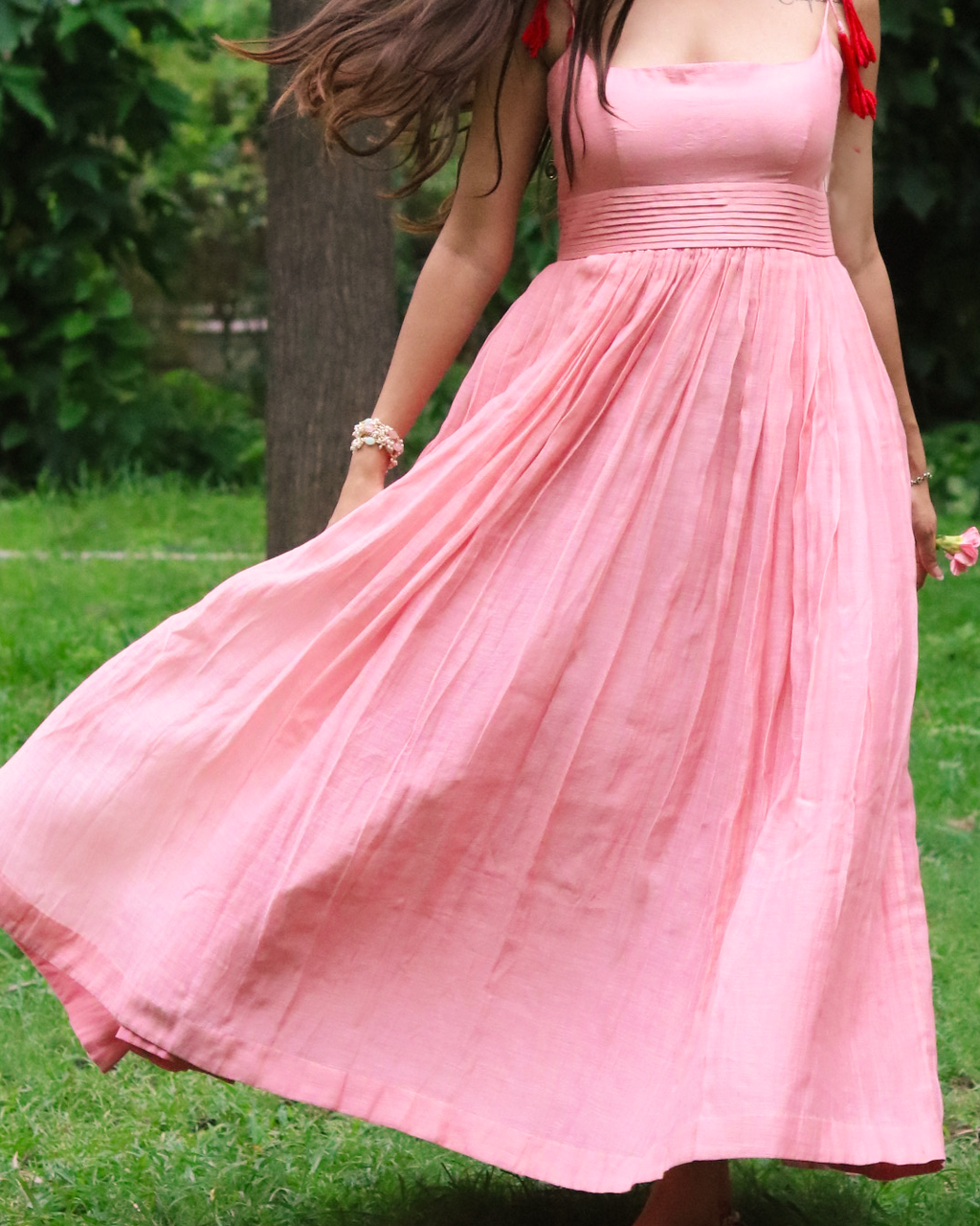 A woman outdoors in a grassy park wearing a flowing pink midi dress with red tassels on the straps. The dress has a square neckline, a gathered elastic waist, and a full skirt that flares out as she moves.