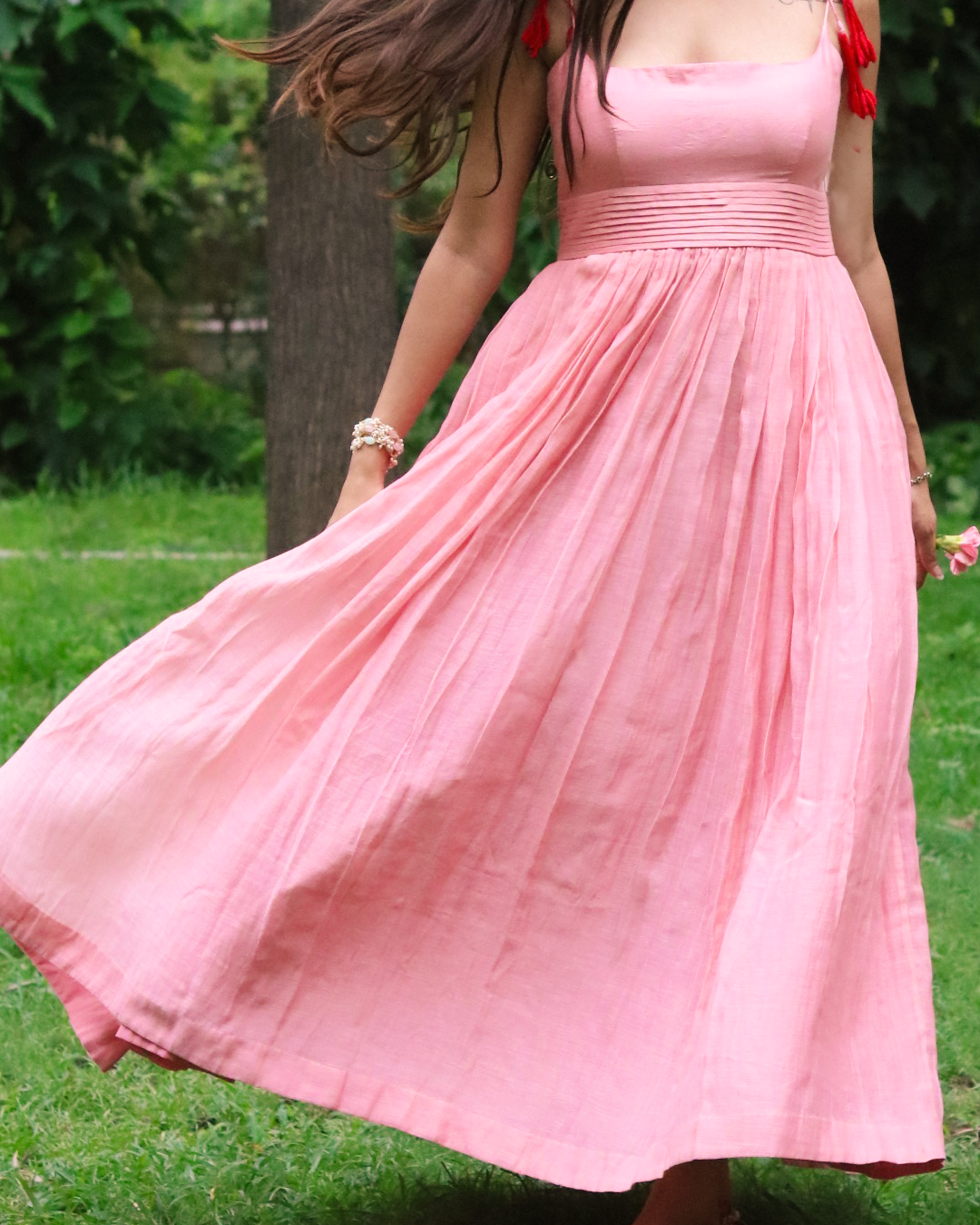 A woman outdoors in a grassy park wearing a flowing pink midi dress with red tassels on the straps. The dress has a square neckline, a gathered elastic waist, and a full skirt that flares out as she moves.