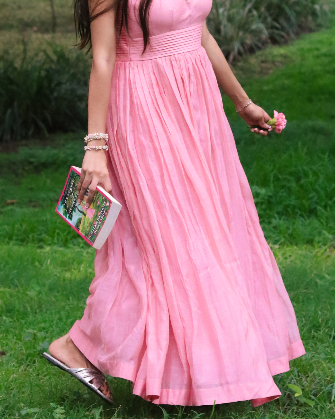 A woman wearing a long pink pleated maxi dress walking outdoors on a grassy lawn, holding a small pink flower and a book.